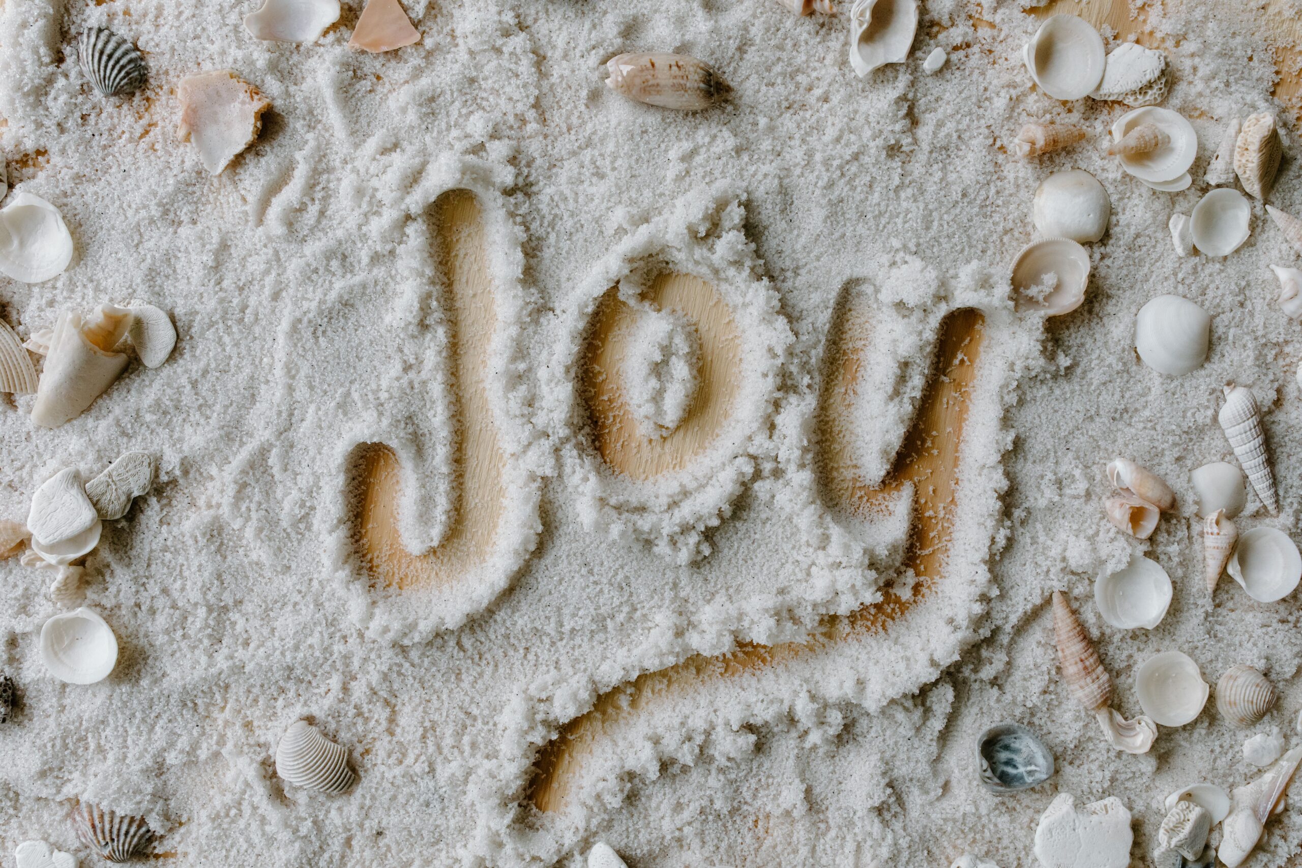 The word 'joy' written in sand with various seashells scattered around.